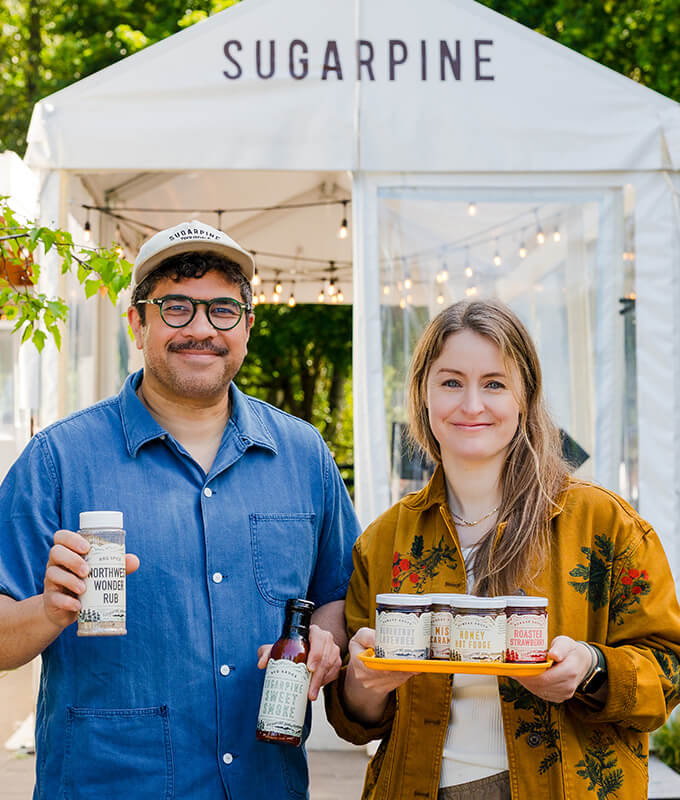 two smiling people holding packaged Sugarpine products outside in front of a tent