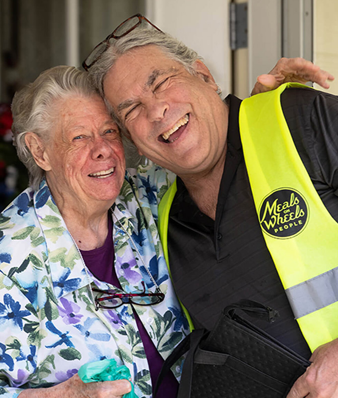 two smiling people hugging, one is wearing a colorful top and the other is wearing a bright yellow vest for Meals on Wheels People
