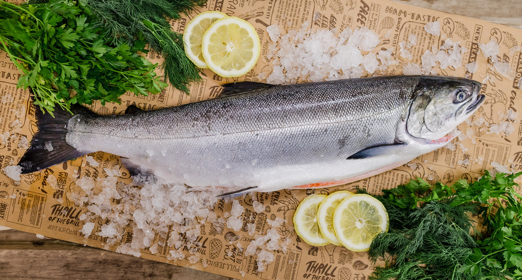 A whole cleaned fish displayed on New Seasons Market brown paper wrap with sliced lemon, fresh herbs, and coarse sea salt on the side.