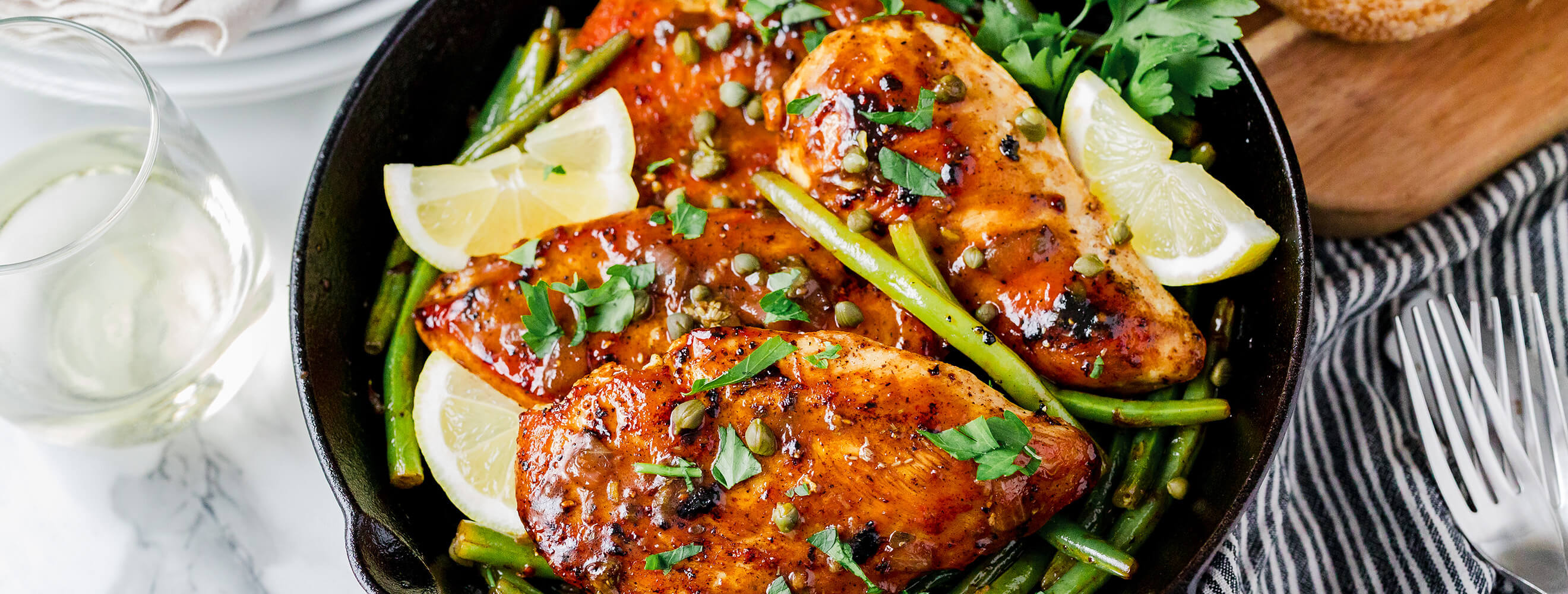 A top down shot of a skillet with cooked Mary's chicken with green beans, lemons, capers and garnished with parsley.