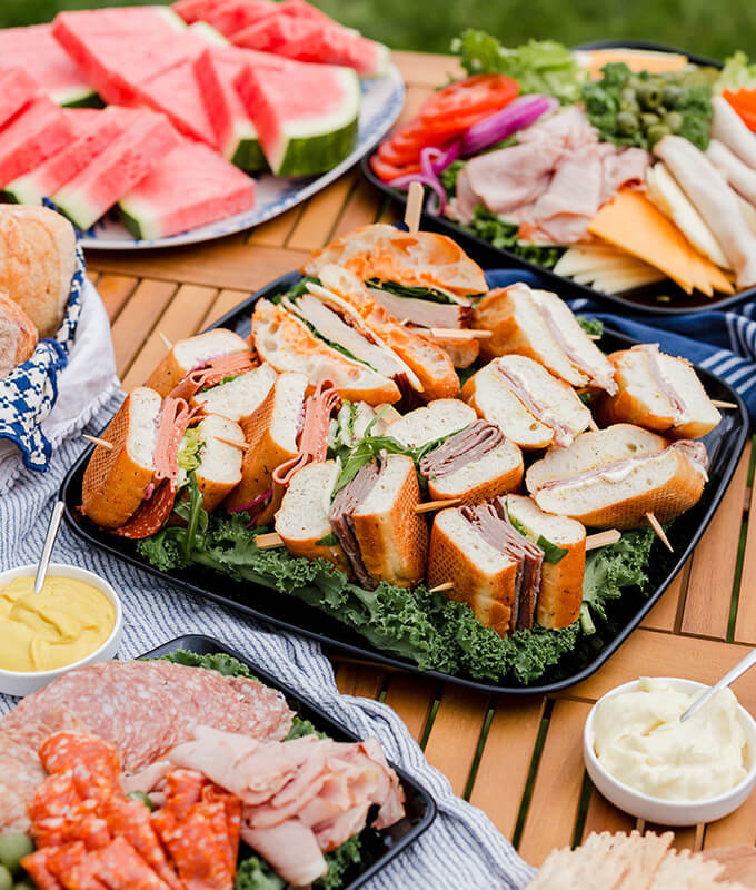 a catering platter with sandwiches on a table with charcuterie and watermelon slices