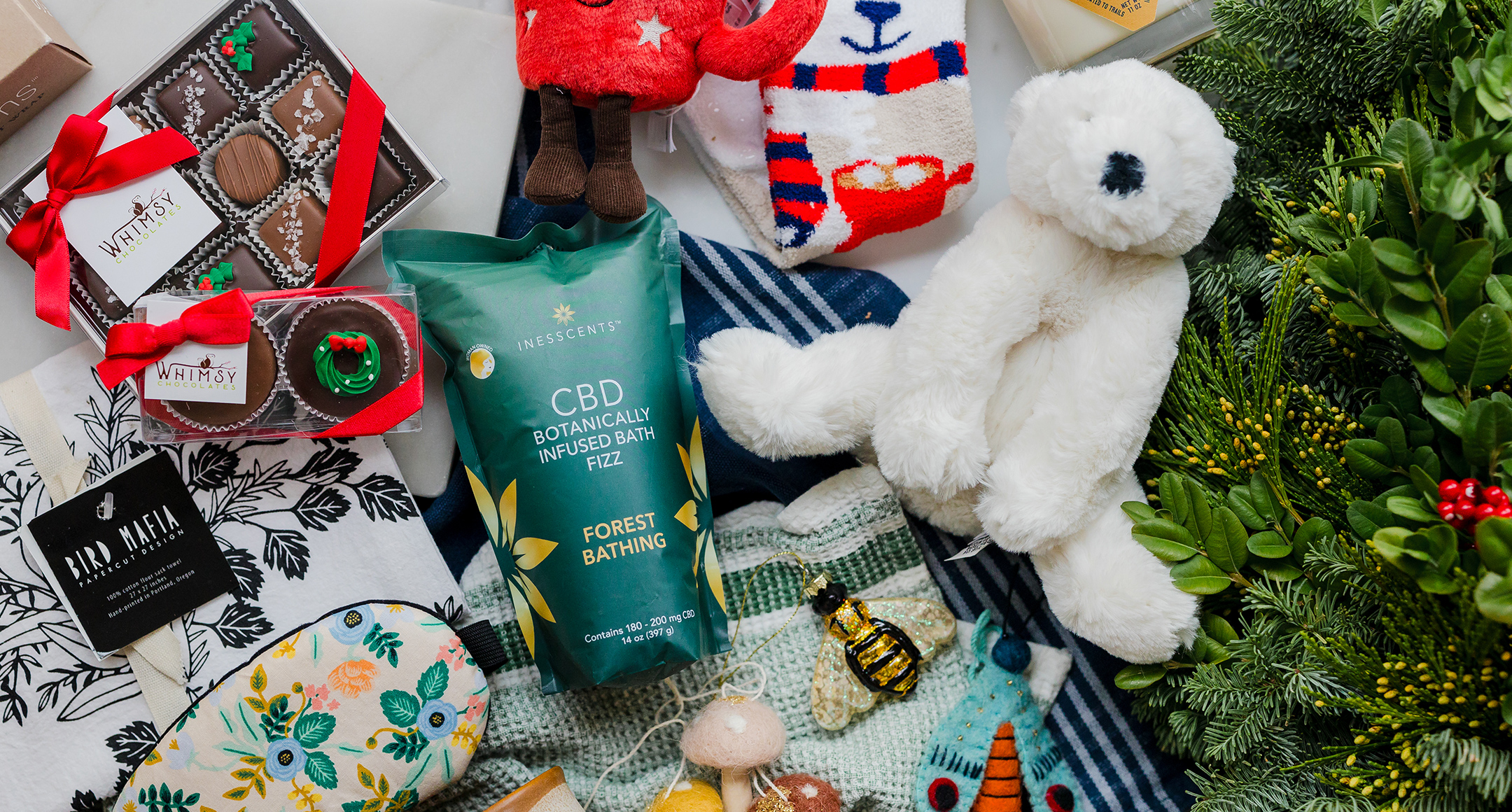 A display of holiday goodies, including a stuffed animal, chocolates, CBD bath salt, tea towels, and tree ornaments.