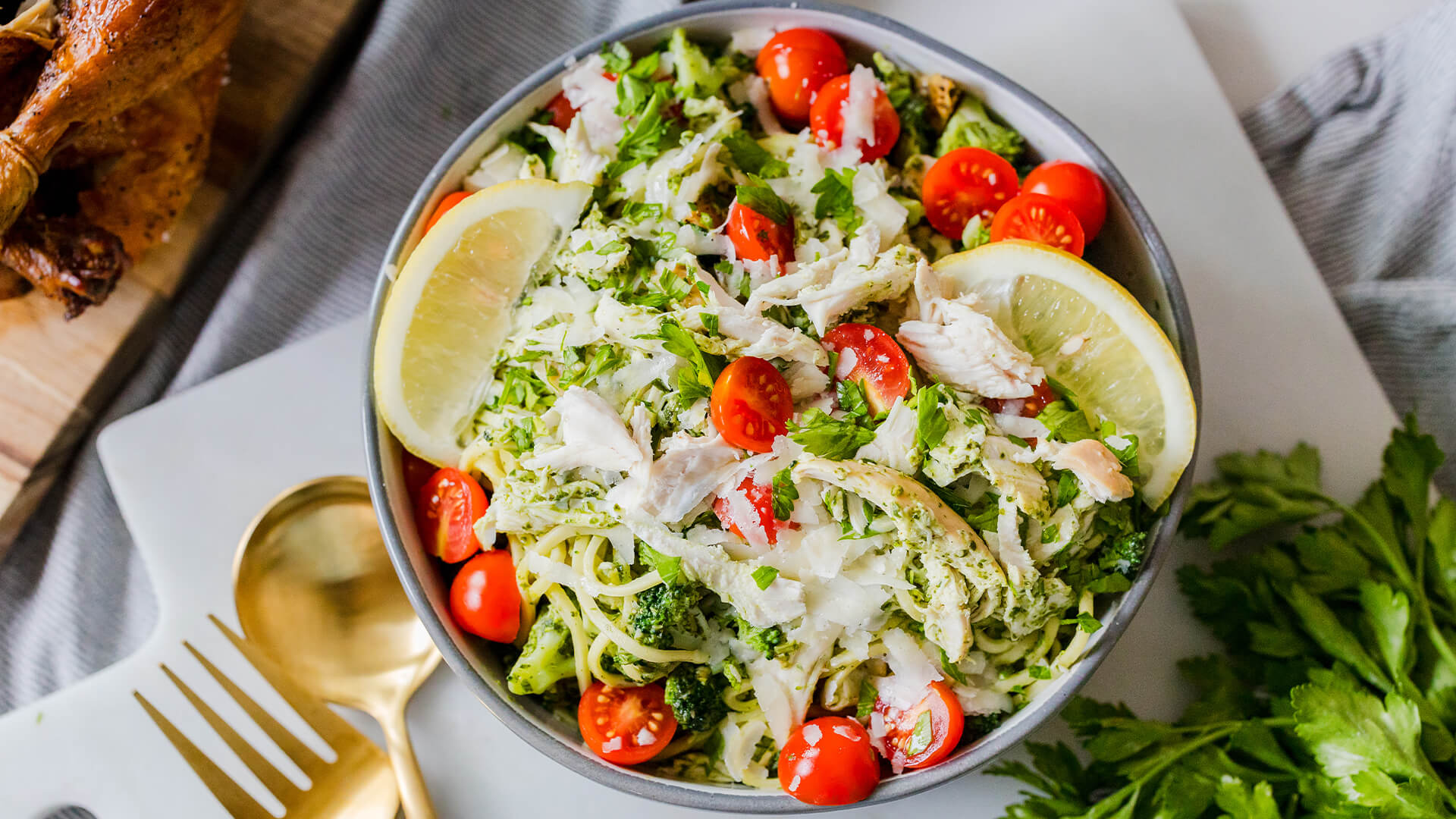 a bowl of chicken pesto pasta sprinkled with cheese, and parsley and topped with lemon wedges and cherry tomatoes.