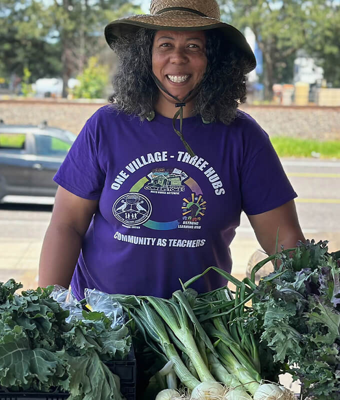 An image of a smiling person in a purple shirt and hat with vegetables in front of them.