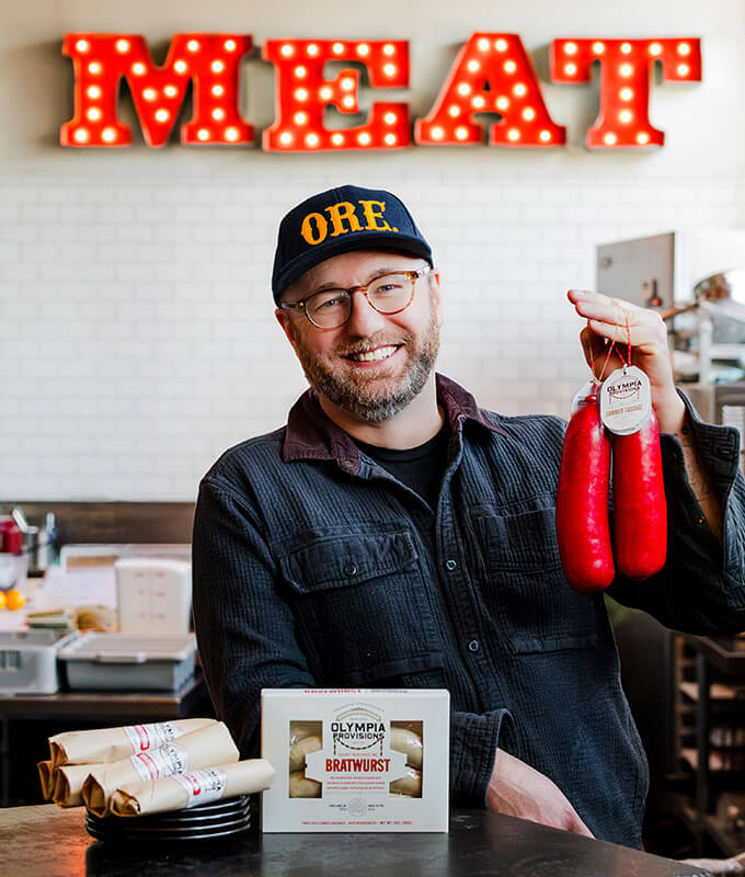 a smiling person in a hat holding Olympia Provisions sausage encased in wax with a sign behind them on the wall that says "MEAT"