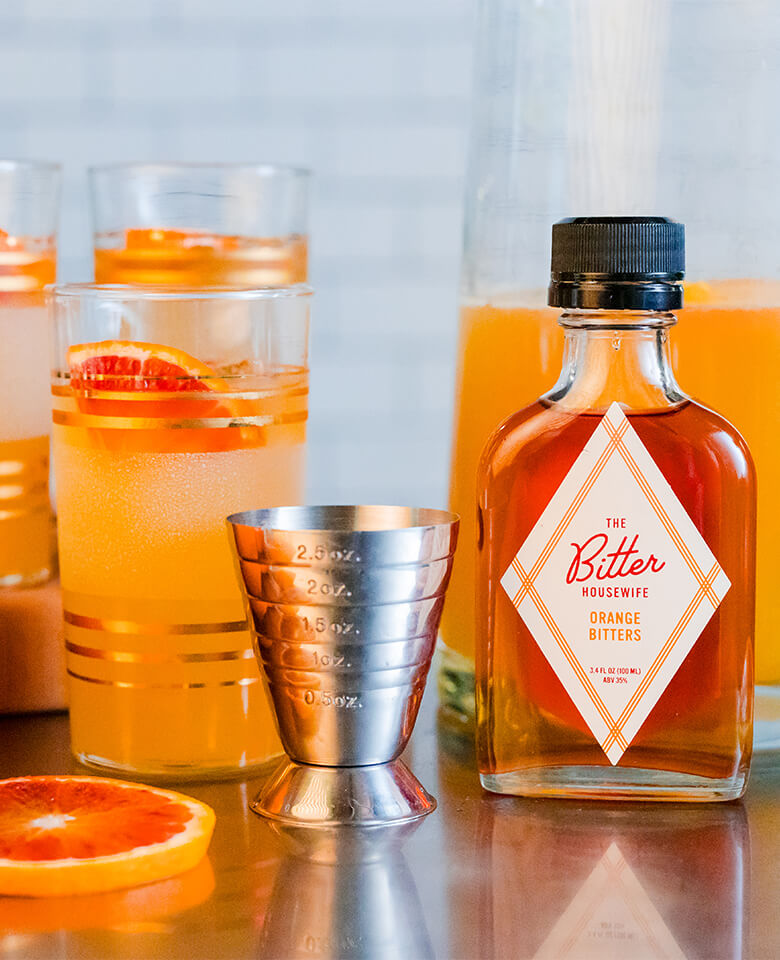 A glass of earl grey spritz garnished with a slice of blood orange placed next to a bottle of orange bitters from the Bitter Housewife.