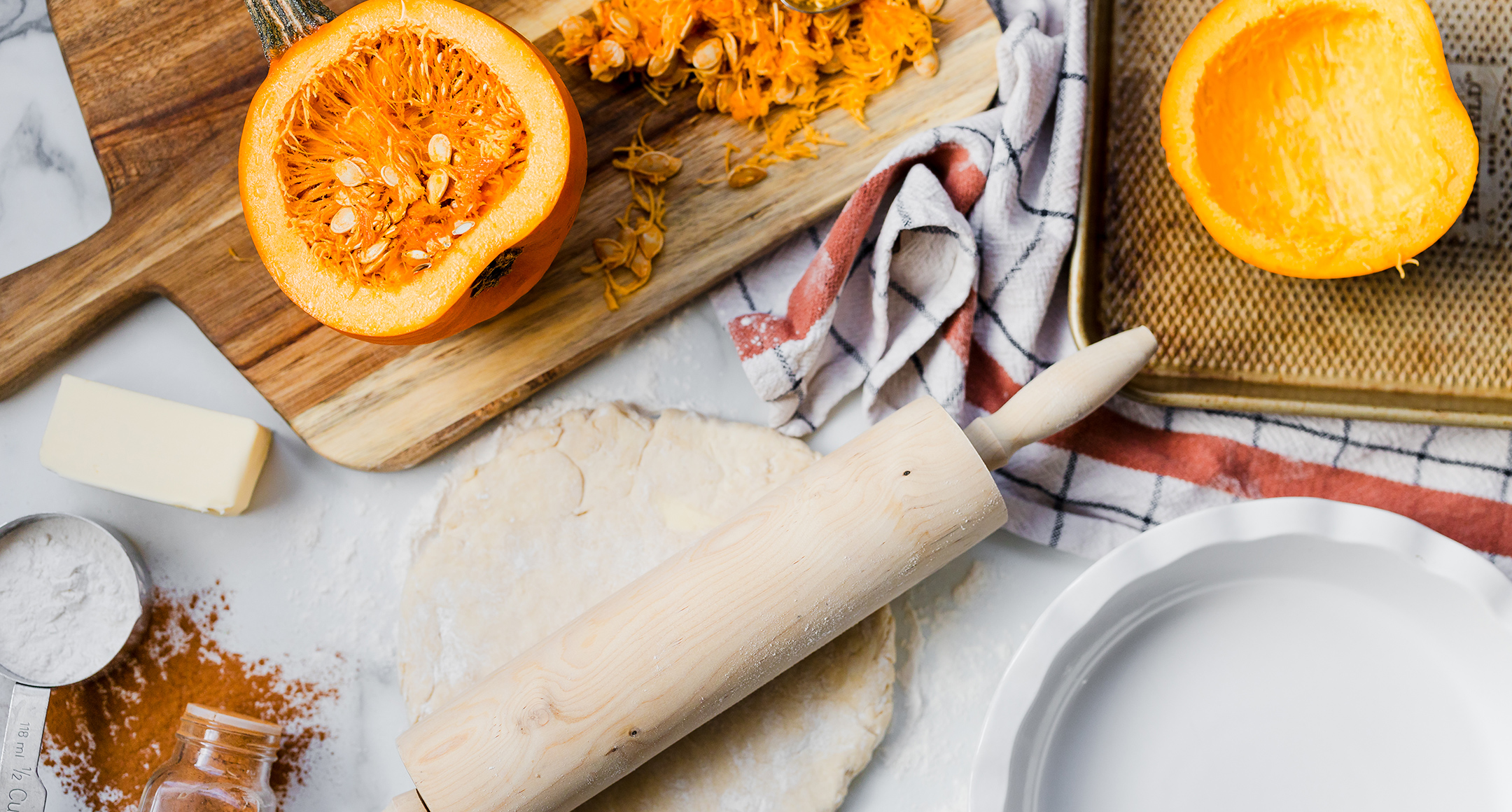 A countertop image with the ingredients of a scratch-made pumpkin pie, including a fresh pumpkin cut open to reveal the insides, spices, and pie dough with a rolling pin. 