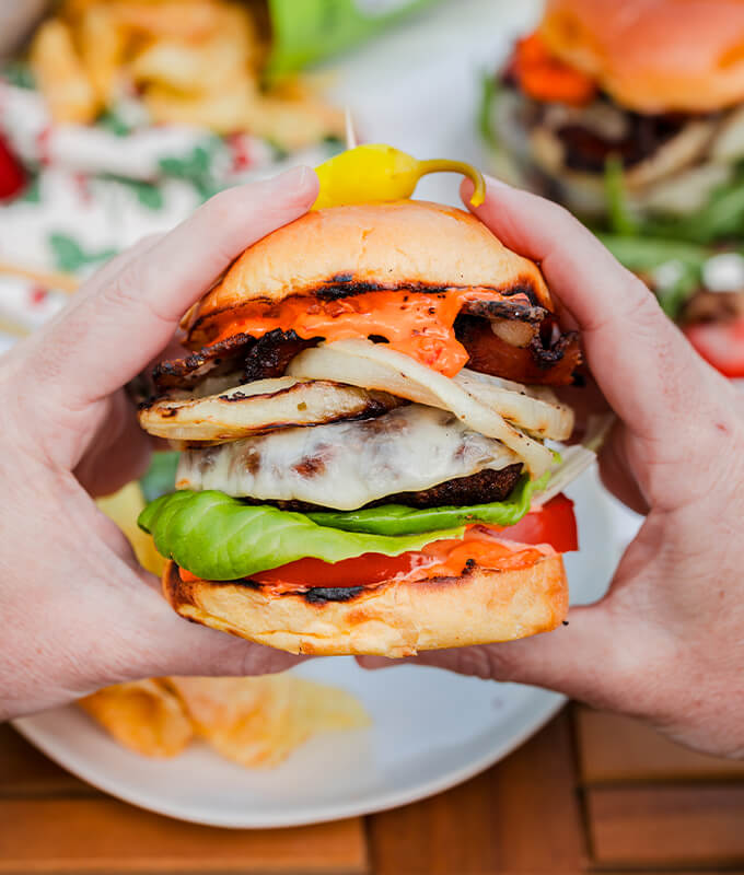 Two hands holding a burger with cheese, tomatoes, grilled onions, lettuce and Calabrian Chili mayo