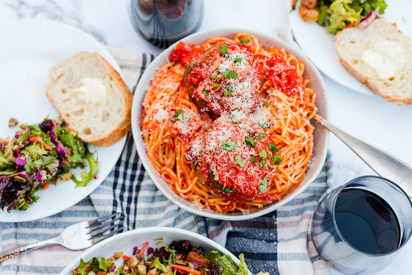 spaghetti and meatballs with freshly grated parmesan on a table with salad, bread and glasses of red wine.