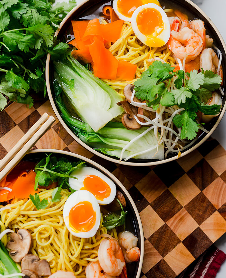 Two bowls of ramen topped with cooked shrimp, bok choy, carrot ribbons, a jammy egg and cilantro