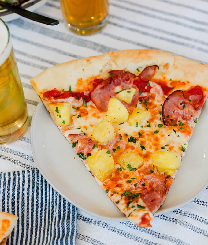 A freshly cooked slice of pizza with ham and pineapple on a white plate on a table with blue and white striped linens.