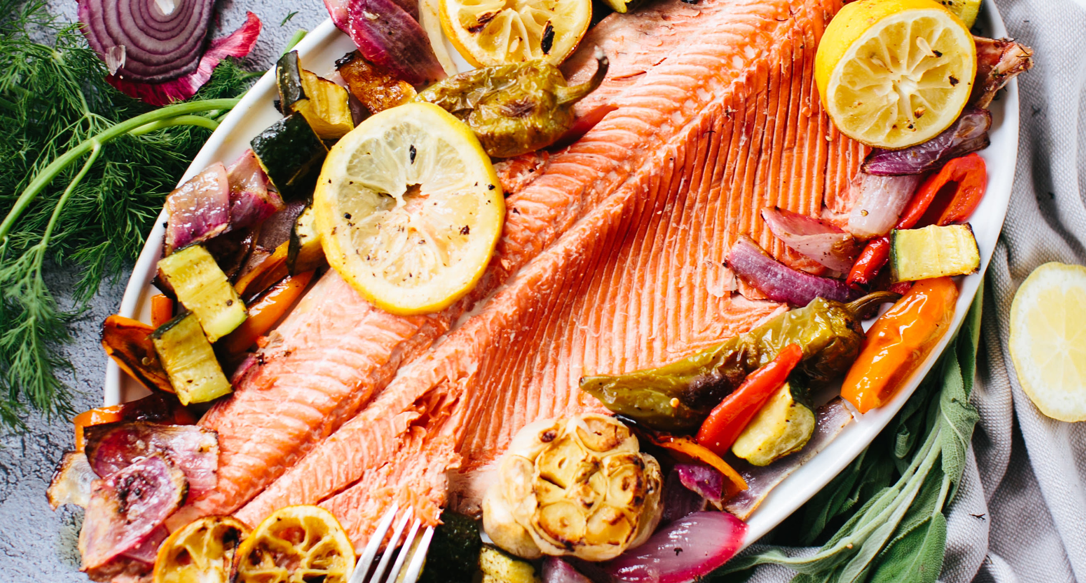 A flaky trout fillet on a platter with a bulb or roasted garlic, sliced lemon, and chopped roasted veggies around the sides.