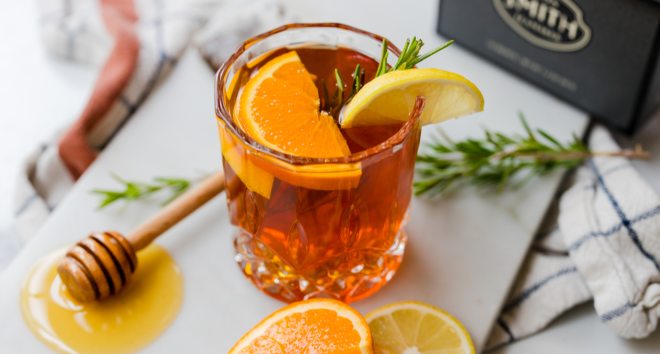 A clear glass of hot toddy garnished with sliced orange, lemon, and a spring of fresh rosemary.