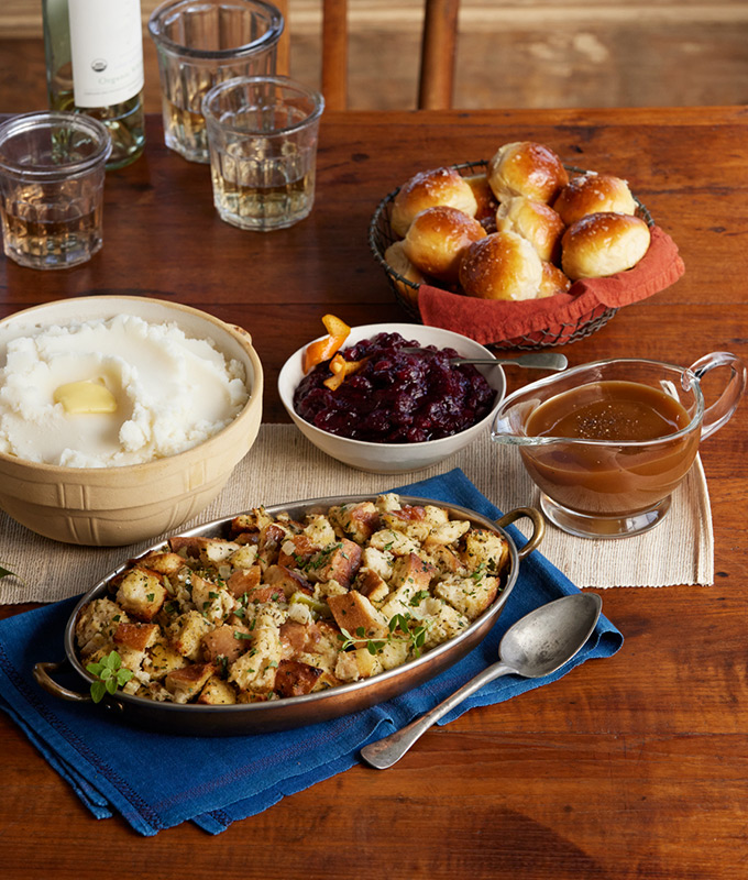 Holiday sides on a festive table including mashed potatoes, cranberry sauce, stuffing, gravy and rolls.