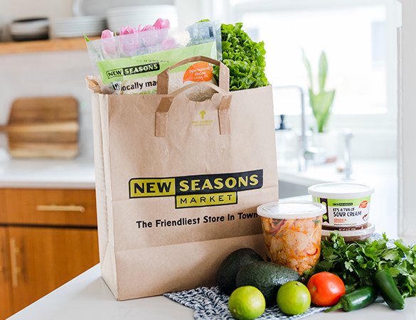New Seasons Market logo paper grocery bag full of groceries and fresh vegetables on a counter